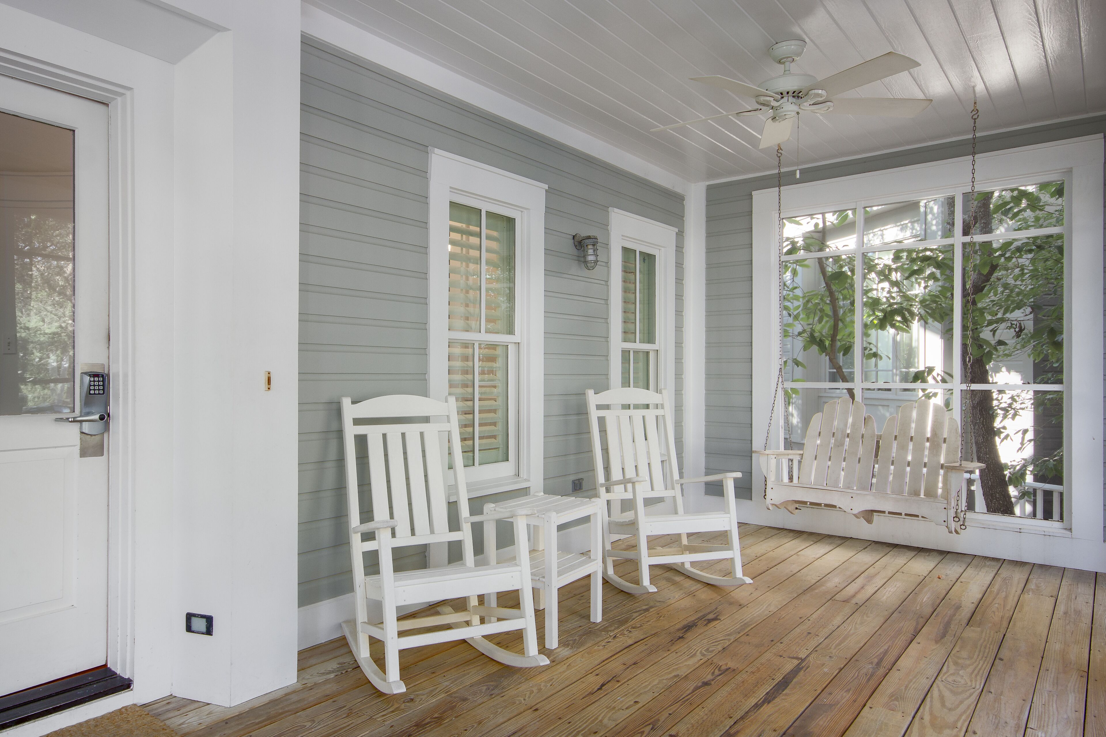 A porch with white rocking chairs and a swing, a door with a keypad lock, and a ceiling fan.