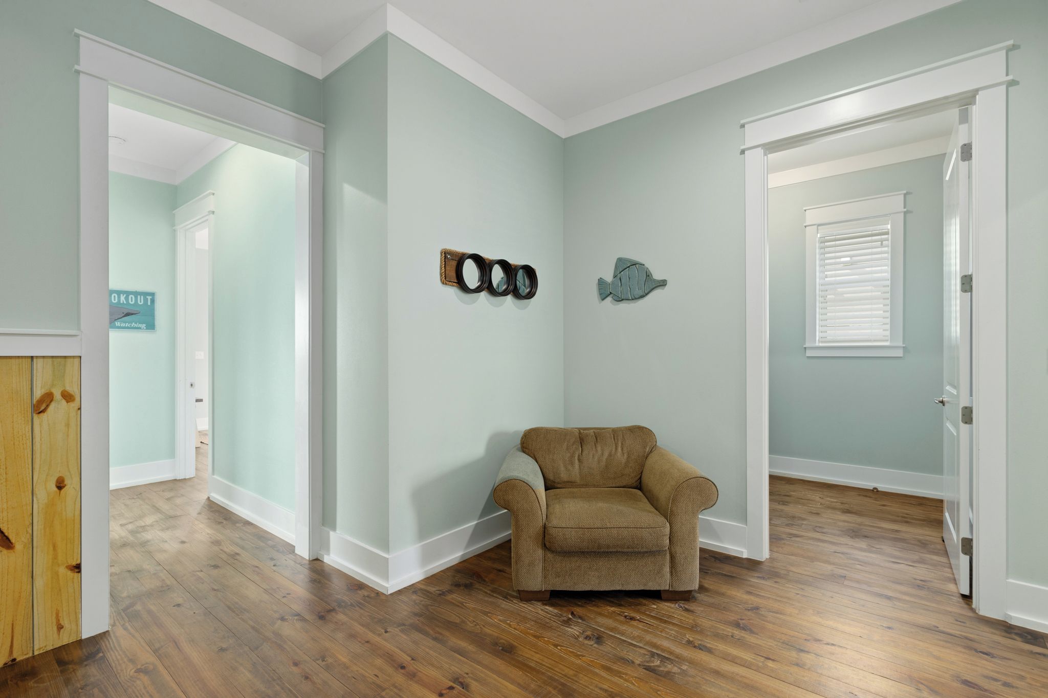 Un pasillo con suelo de madera, un sillón beige y decoraciones en la pared, incluyendo un par de gafas y un pez.