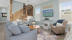 A living room with a grey sofa, a beige armchair, a wooden staircase, a fish wall art, and a TV on a white shelf.