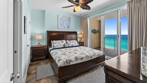 A bedroom with a bed, a ceiling fan, a nightstand, a lamp, a window with a view of the ocean, and a door. - Luxurious Ocean Villa Resort - Gulf Front Paradise With Views! (Panama City Beach)