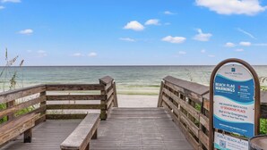 Un muelle de madera que conduce a una playa, con un letrero que indica avisos de banderas y datos sobre el acceso.