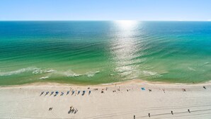 Une vue aérienne d’une plage avec une eau d’un bleu limpide, un rivage sablonneux, ainsi que plusieurs parasols et des personnes.