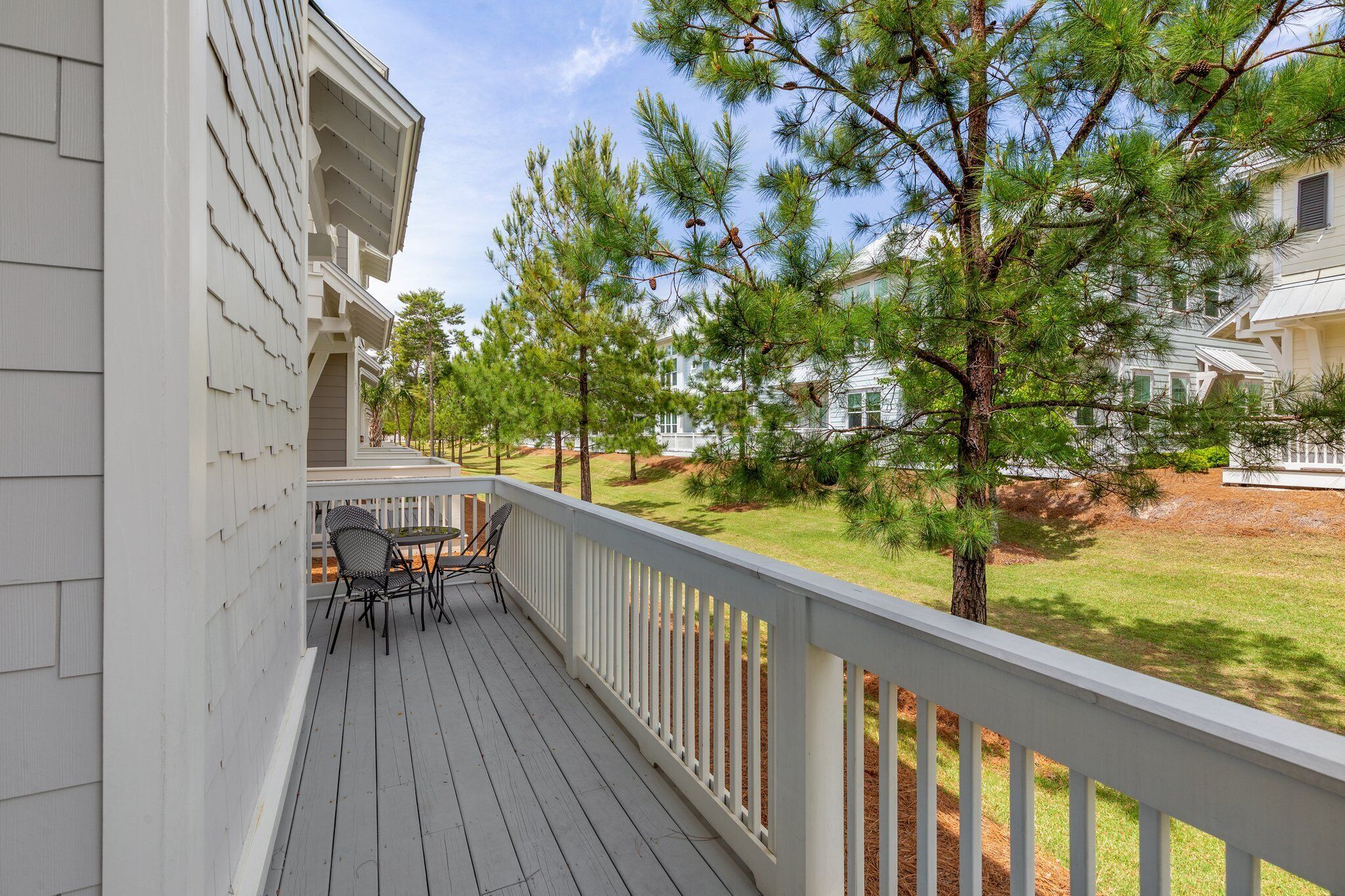 A wooden deck with a white railing, a table with chairs, and pine trees in the background.