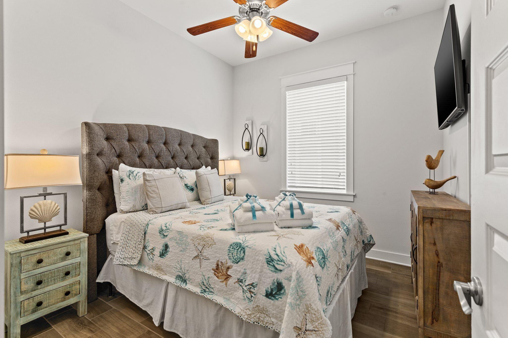 Une chambre à coucher avec une tête de lit capitonnée, un lit avec une literie à motifs floraux, une table de chevet avec une lampe, une commode en bois et un ventilateur de plafond.