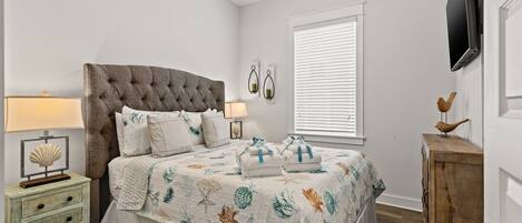 Une chambre à coucher avec une tête de lit capitonnée, un lit avec une literie à motifs floraux, une table de chevet avec une lampe, une commode en bois et un ventilateur de plafond.