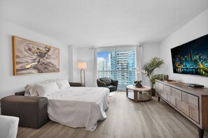 A modern hotel room with a large bed, a seating area with a chair and table, a TV, and a cityscape view.