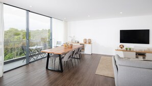 Ein modernes Wohnzimmer mit Essbereich, einer Couch, einem Fernseher und einem Balkon mit Blick auf Bäume.