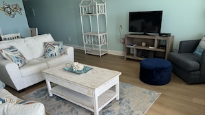 Un soggiorno con un divano grigio chiaro, un tavolino da caffè, un poggiapiedi blu, un mobile TV e una mensola decorativa in metallo.