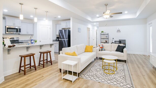 Cocina moderna con armarios blancos, microondas y refrigerador. Hay un sofá blanco con almohadones amarillos y una mesita blanca. El cuarto tiene piso de madera y un ventilador de techo.