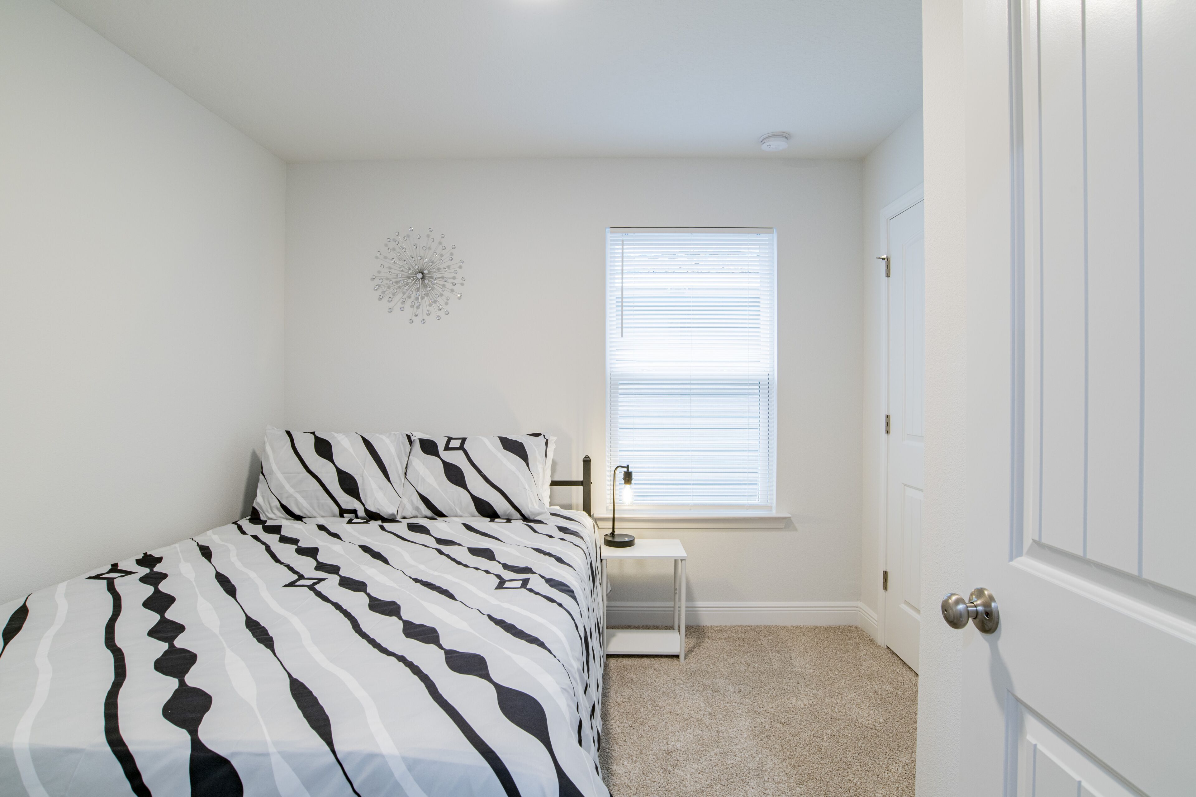 A bedroom with a bed, a bedside table, a lamp, a window with blinds, and a door.