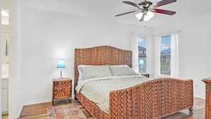 Une chambre à coucher avec un lit à motifs tissés, une table de chevet en bois avec une lampe, un ventilateur de plafond et une vue sur l’extérieur grâce à de grandes fenêtres.