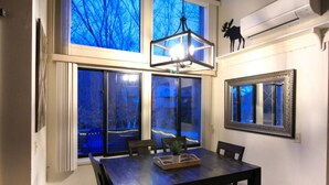 A dining area with a dark wooden table, four chairs, a hanging light fixture, and large windows with blue-tinted glass. - Loon Outlook - Best View, <3min to Loon (Lincoln)