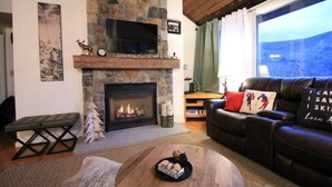 A cozy living room with a stone fireplace, a leather sofa, and a large window offering a view of the outdoors. - Loon Outlook - Best View, <3min to Loon (Lincoln)