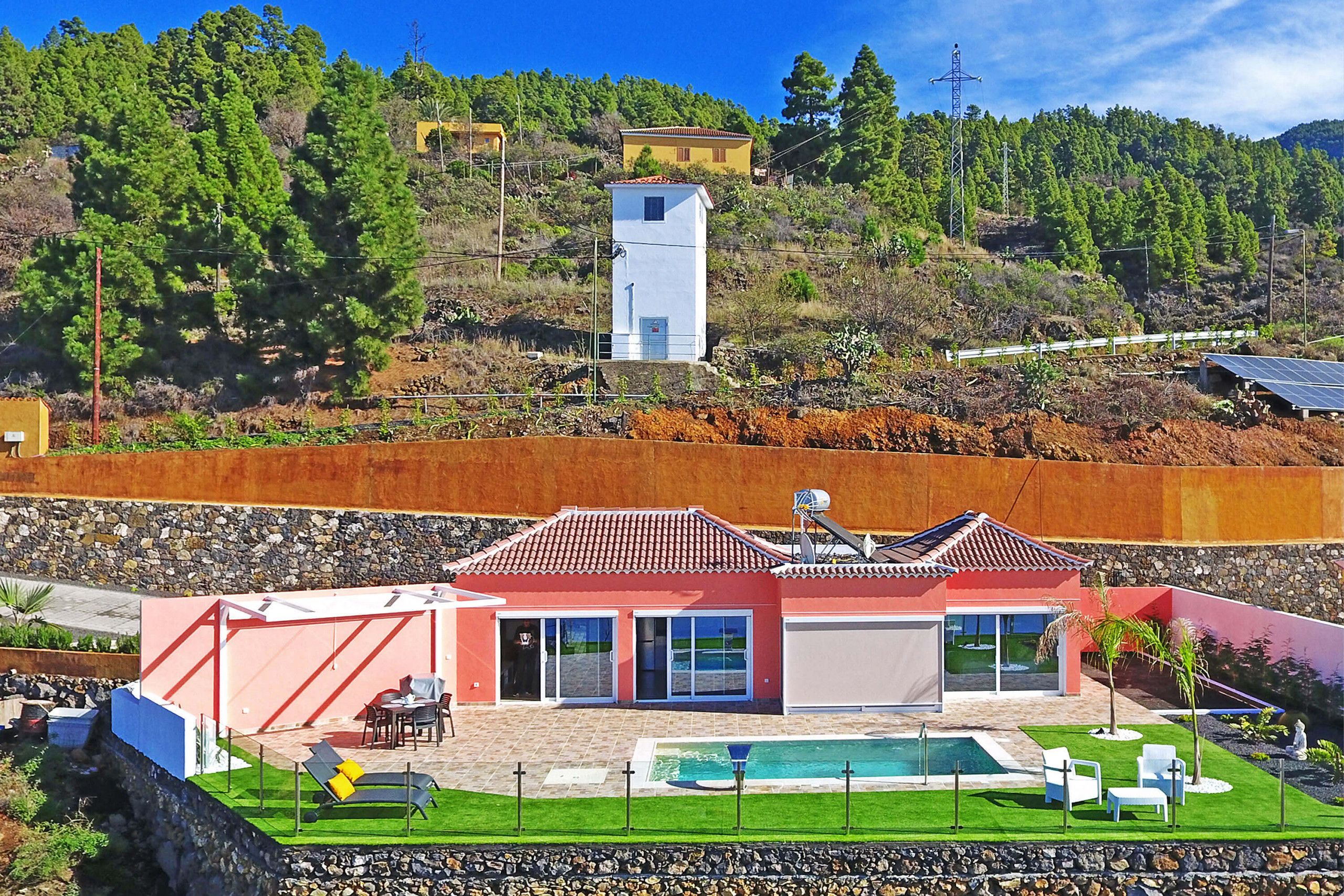 A modern house with a swimming pool, a red roof, and a view of a hill with trees.