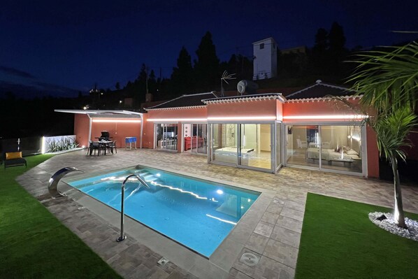 A swimming pool with a glass enclosure, a patio area with outdoor furniture, and a covered seating area.