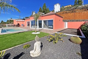 Ein modernes Haus mit Schwimmbad, einer Terrasse und Gartenmöbeln sowie einem gepflegten Garten mit Statue und Kakteen.