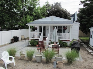 A small white house with a porch, outdoor seating, and potted plants.