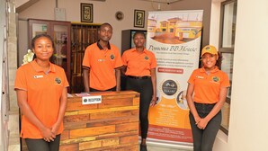 Four individuals in orange uniforms standing in front of a reception desk with a wooden backdrop and a promotional poster for Pomonabbhouse.