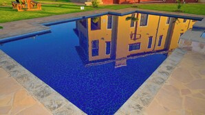 A rectangular swimming pool with a clear blue water surface, surrounded by stone tiles and a grassy area with a building reflection.