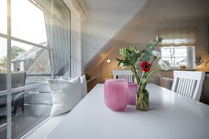 Un comedor con una mesa blanca, un jarrón rosa con flores y un florero de vidrio con follaje verde.