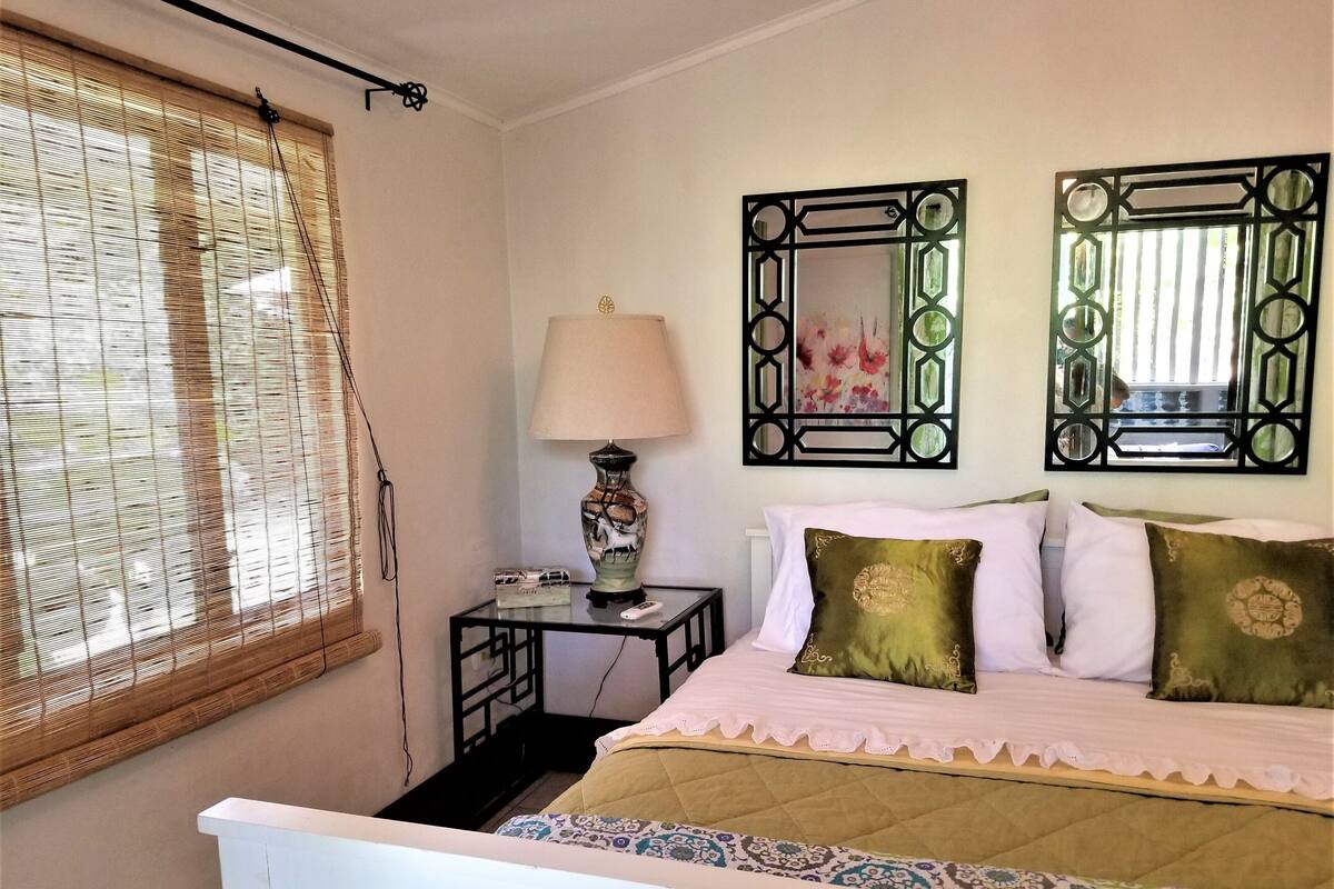 A bedroom with a bed, a bedside table with a lamp, a mirror, and a window with blinds.