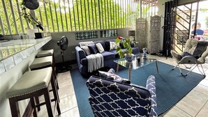 A cozy living area with a sofa, a coffee table, and a chair. There is a window with vertical blinds, a decorative ceiling fan, and a view of greenery outside. - Casa Sophia (Guapiles)