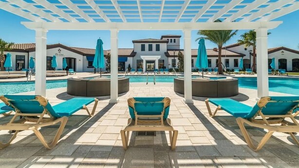 A pool area with sun loungers and umbrellas, a pergola overhead, and a building with a tiled roof in the background. - Paradise 6 BR home - private pool and spa near all Disney parks - Free pool heat (Kissimmee)