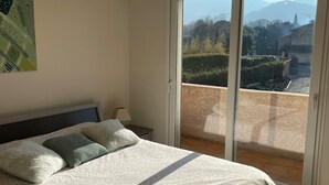 A bedroom with a bed, pillows, a nightstand, a lamp, a ceiling light fixture, and a sliding glass door leading to a balcony with a view of the outdoors.
