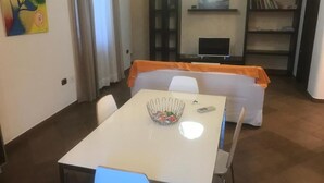 Un comedor con una mesa y sillas blancas, un cuenco de fruta y un televisor al fondo.