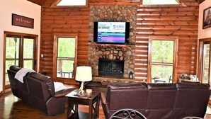 A log cabin interior with a stone fireplace, leather sofas, a television, and a 'welcome' sign.