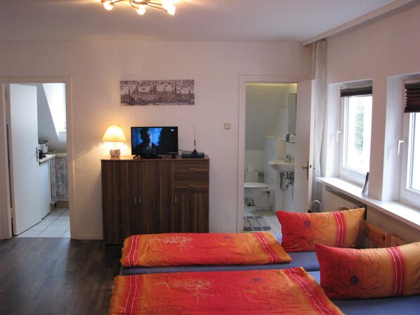Ein Schlafzimmer mit Fernseher, einem Bett mit roter und blauer Bettwäsche, einer Holzkommode und einem durchgegangenen Tür erkennbaren Badezimmer.