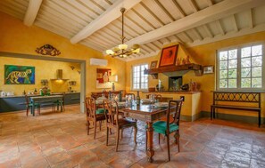 A dining area with a wooden table and chairs, a chandelier, and a kitchen in the background.