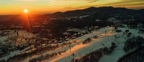 A snow-covered ski resort with a sun setting in the background, ski lifts, and a mountain landscape.