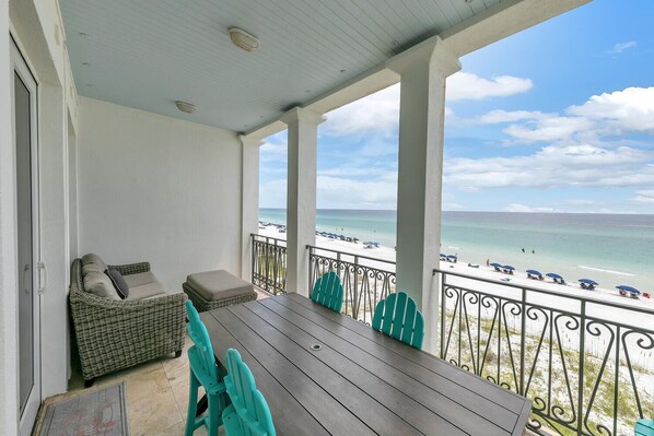 A covered balcony with wicker furniture, a table, and chairs, overlooking a beach with umbrellas and people. - Remodeled Beachfront😎😎 Frangista Delos⛱️⛱️Heated Splash🐠🐠Pool Elevator🐬🐬Beach Serv (Miramar Beach)