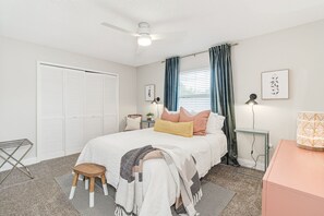 Un dormitorio con cama, mesita de noche, lámpara, silla, taburete pequeño, armario y ventana con cortinas.