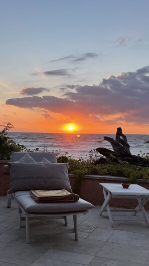Une vue sur le coucher de soleil depuis une terrasse agrémentée d’un banc et d’une table, d’où l’on aperçoit la mer et une formation rocheuse remarquable.