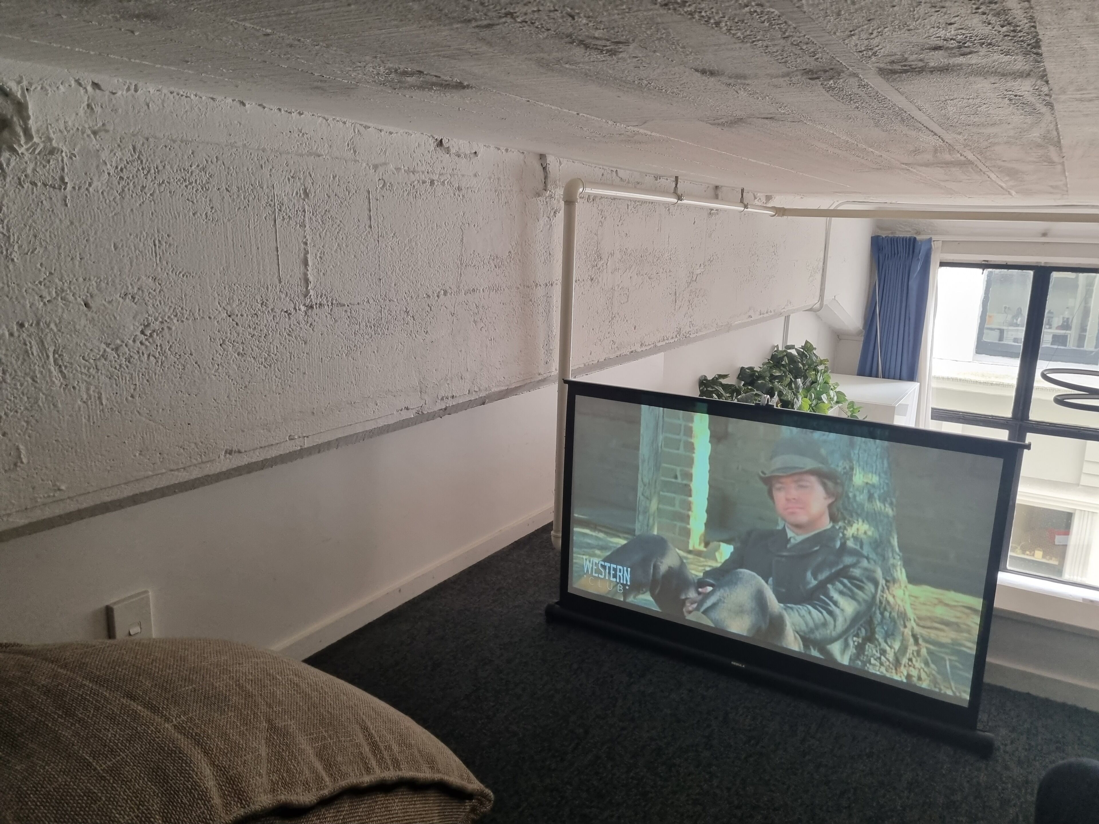 Quarto com uma televisão de tela grande na parede, uma janela com cortinas azuis e uma planta no parapeito.