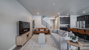 Una sala de estar moderna con sofá, reposapiés y televisor. Una escalera conduce a una zona de cocina con armarios oscuros y un refrigerador.
