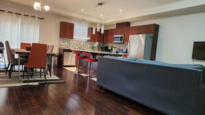 A modern kitchen with wooden cabinets, a built-in microwave, and a refrigerator. There is a dining area with a table and chairs, and a sofa in the living area. - Beautiful Three Bedroom House (Kitchener)