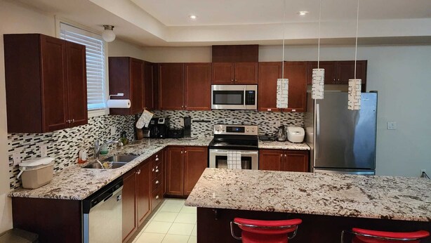 A modern kitchen with dark wooden cabinets, a granite countertop, stainless steel appliances, and a central island with red barstools. - Beautiful Three Bedroom House (Kitchener)