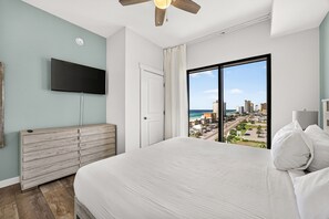 Ein Schlafzimmer mit einem großen Bett, einem Flachbildfernseher, einer hölzernen Kommode und Blick auf eine Stadtansicht durch ein großes Fenster.