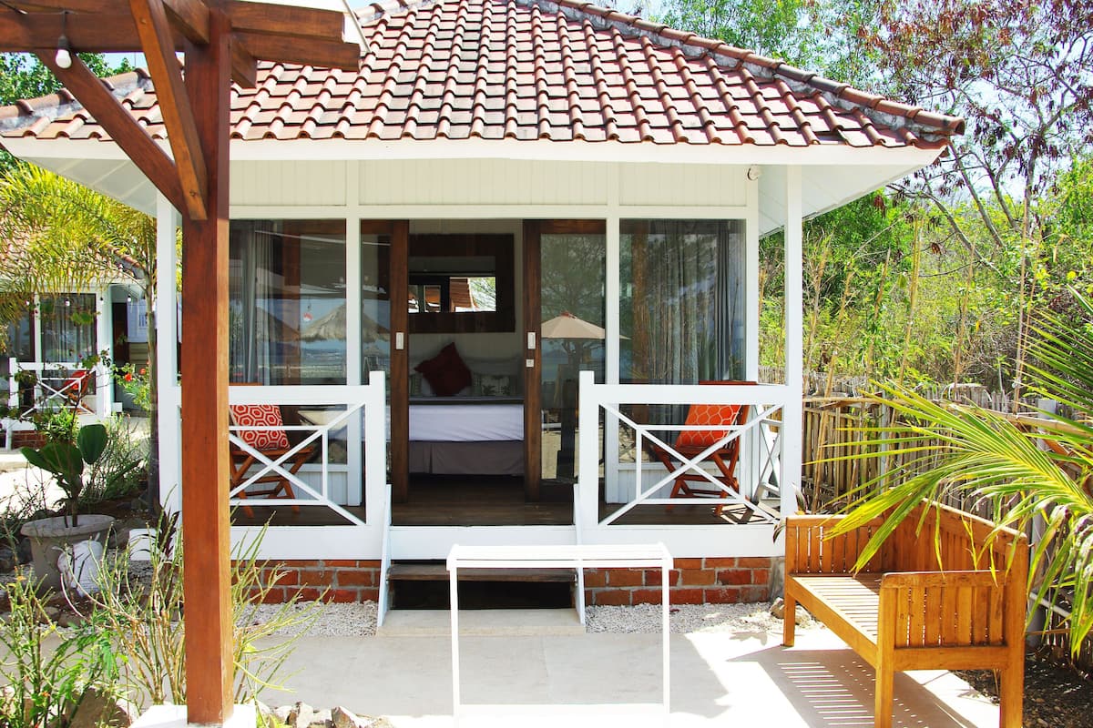 A small, single-story house with a tiled roof, a balcony with a wooden bench, and a garden area with plants and a palm tree.
