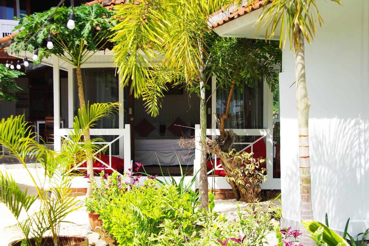A covered outdoor seating area with a bed, surrounded by lush greenery and plants.