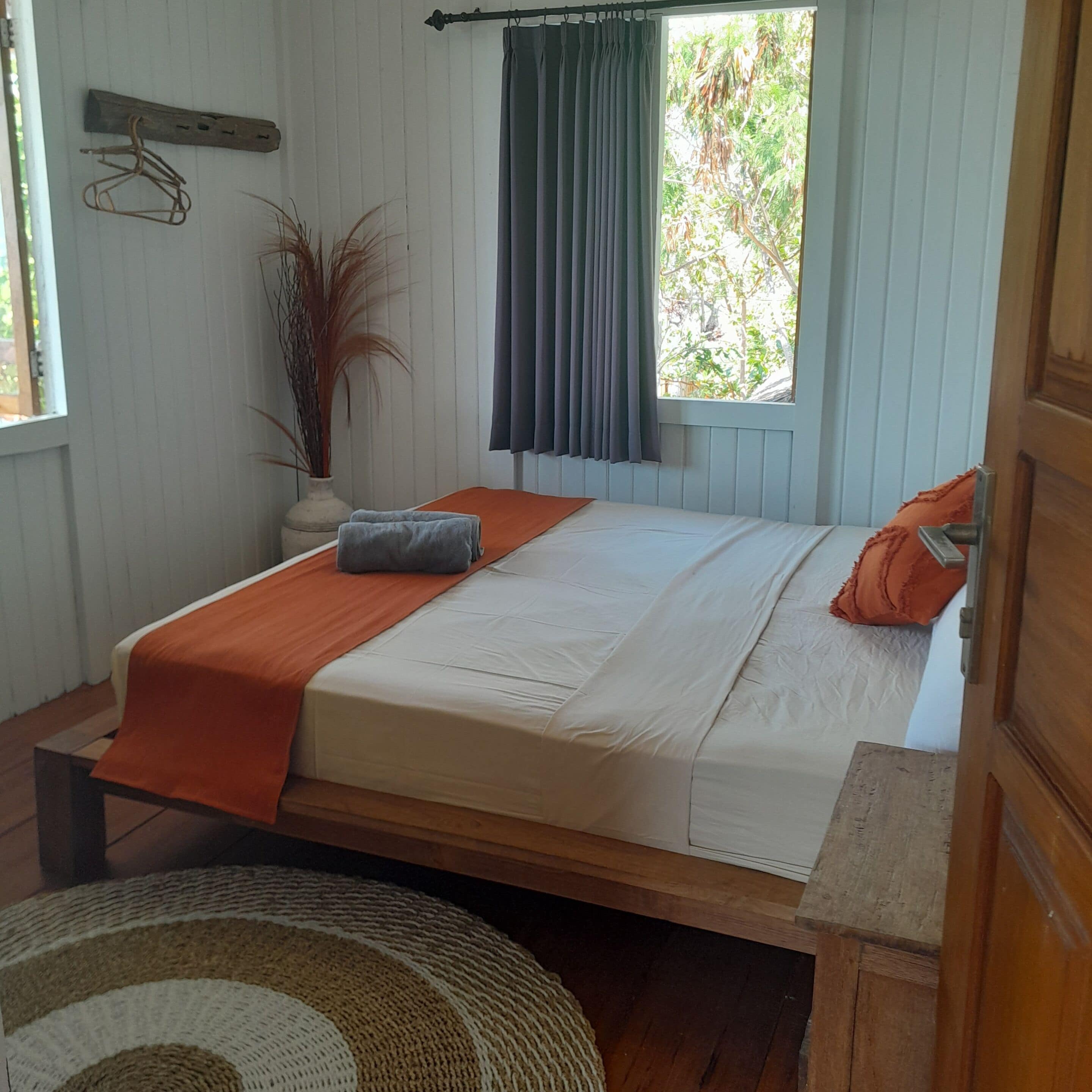 Un lit simple avec un cadre en bois, un oreiller gris et une couverture orange. Une petite plante en pot est posée sur la table de chevet, à côté du lit.