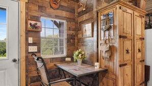 A rustic kitchen with wooden walls, a dining table with chairs, a window with blinds, and a cabinet with decorative items.