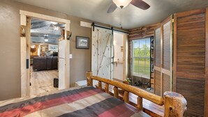 A bedroom with a bed, wooden headboard, a ceiling fan, and a view of the kitchen through an open door.