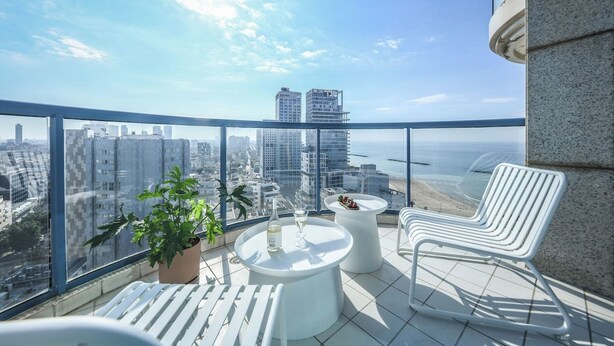 A balcony with a city view, two potted plants, a round table with a bottle and glass, and two white chairs. - Beachside 2BR Apt with Sea & Skyline Views (Tel Aviv-Yafo)