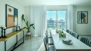A modern dining area with a long table, chairs, and a view of the city through large windows. - Beachside 2BR Apt with Sea & Skyline Views (Tel Aviv-Yafo)