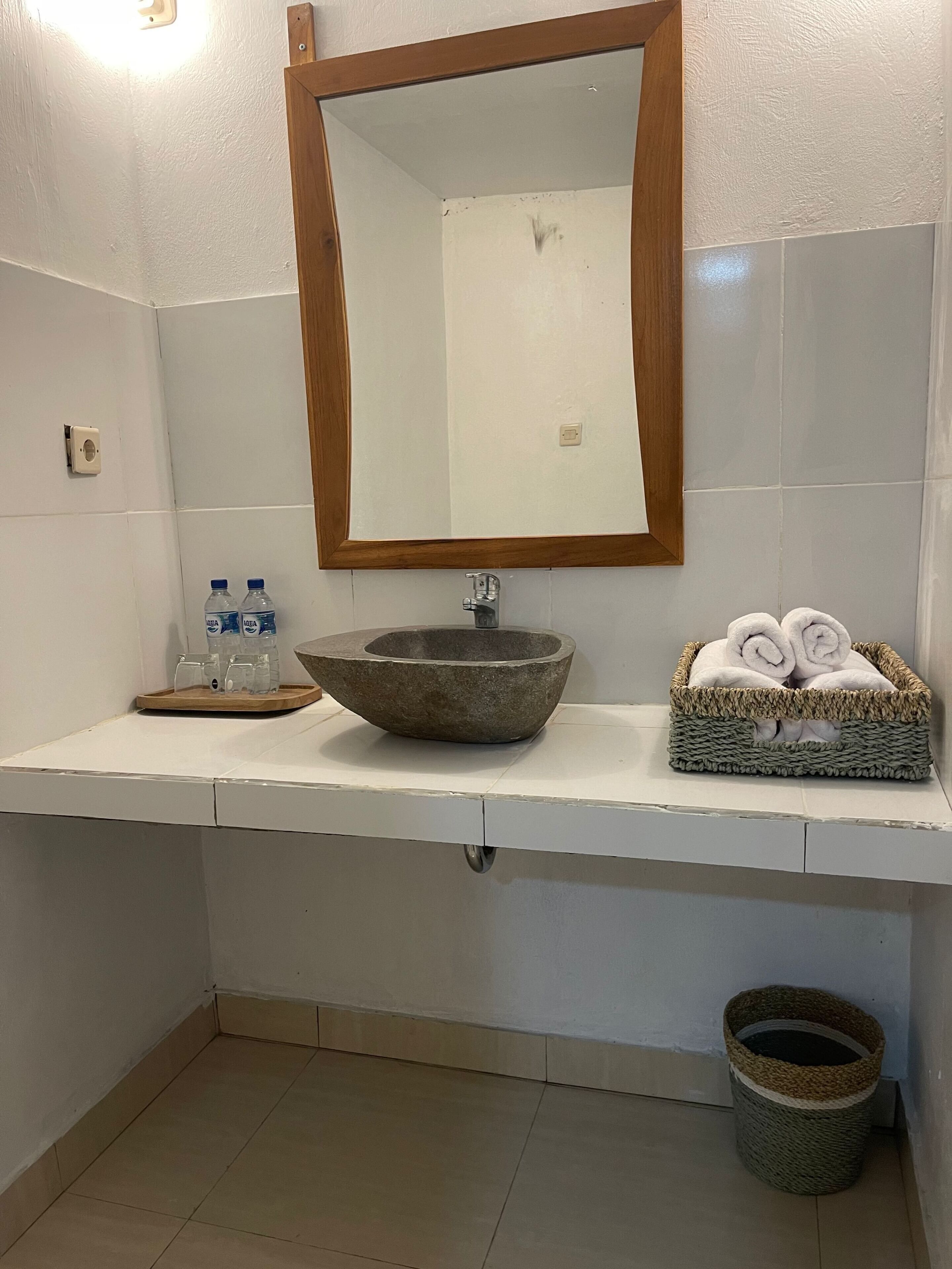 A bathroom with a stone basin sink, a wooden-framed mirror, a tray with water bottles, and a basket of towels.
