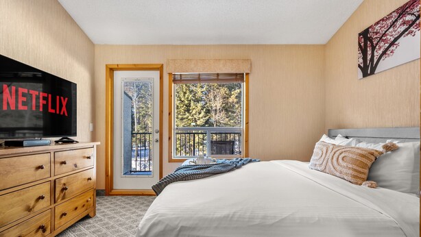 Un dormitorio con cama, televisor en una cómoda, ventana con vista a árboles y una puerta.